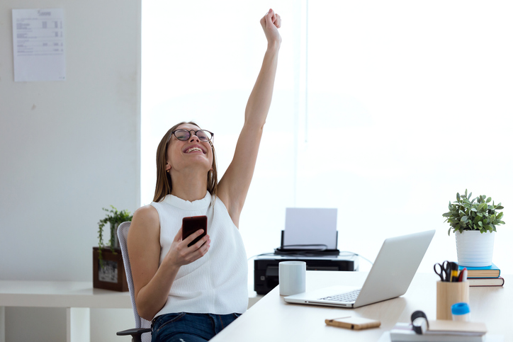 Woman at desk with phone in hand with one arm up celebrating